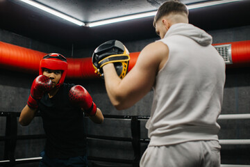 A teenager wearing a red protective helmet and gloves trains in a boxing ring with a coach.