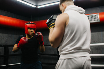 A teenager wearing a red protective helmet and gloves trains in a boxing ring with a coach.