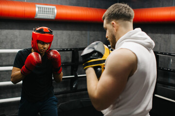 A teenager wearing a red protective helmet and gloves trains in a boxing ring with a coach.