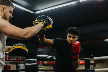Young man, dressed in black t-shirt and red boxing gloves, boxing in the ring with her coach..