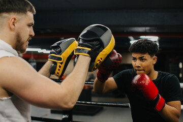 A male trainer helps a teenage boxer with striking techniques in a boxing ring.