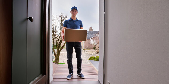Smiling delivery worker, delivering a package to a home, standing at the open front door holding a cardboard box.