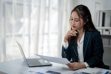 A woman in a business suit is sitting at a desk with a laptop and a piece of paper. She is focused...