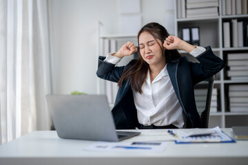 A woman is sitting at a desk with a laptop and a stack of papers. She is wearing a black jacket and she is in a state of exhaustion