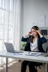 A woman sits at a desk with a laptop and a stack of papers. She is wearing a black suit and she is in a state of stress or frustration