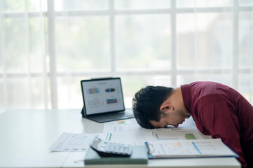 A man in a red shirt is slumped over a desk with a laptop and a stack of papers