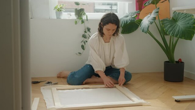 Young woman stretches canvas onto frame, fixing fabric to wooden frame with construction stapler. Preparing canvas for painting.Pleasant pastime for hobby on sunny weekend day at home. Creativity, DIY