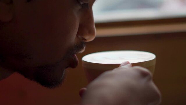 Closeup shot of Indian man drinking coffee from coffee cup while sitting inside a cafe. Man taking a sip of coffee from the cup. Man enjoying his hot coffee inside the cafe. Morning breakfast in caf&eacute;.