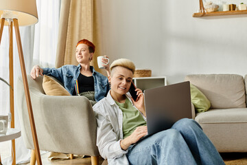 A lesbian couple relaxes on a couch, sipping coffee and using a laptop together.