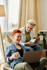 Two women with short hair sit on a couch, engrossed in a laptop screen, sharing a moment of connection.