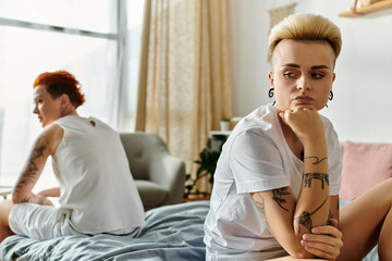 Two women with tattoos, a lesbian couple, sit together on a bed in a bedroom, showcasing...