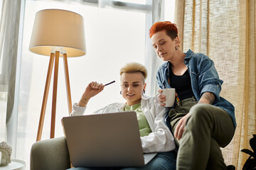 Two women, a lesbian couple, sit on a couch looking at a laptop together, engrossed in the screen...