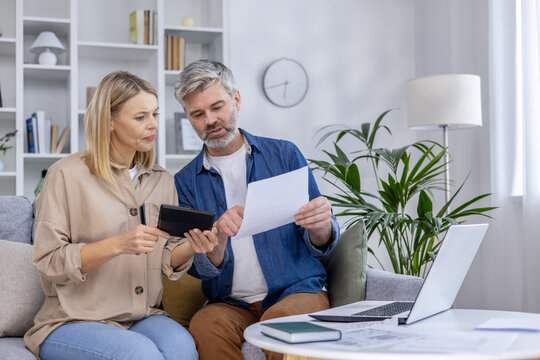 Mature couple sitting on a couch, reviewing documents and using a tablet at home. They seem focused and engaged in a financial discussion.