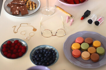 Plate of pastel macarons, cookies and chocolate, cup of tea of coffee, glass of bubble water, various berries, books and accessories on the table. Selective focus, pastel colors.

