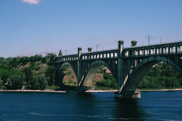 Vintage arched bridge crossing over a river, featuring railway tracks on top and surrounded by lush greenery with a hint of urban buildings in the background
