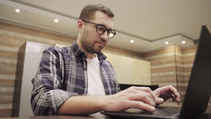 Young man in glasses working on laptop in modern kitchen. Slow motion - Powered by Adobe