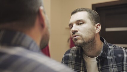 Young man touching his beard, checking, standing in bathroom, looking at mirror