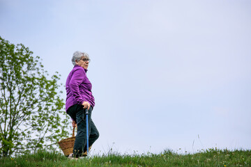 An elderly woman with short gray hair walks up a grassy hill with a basket.