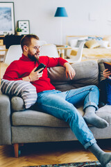 Couple in love resting at apartment on weekends, male using mobile phone and wireless internet at home while his girlfriend reading interesting literature,family recreating at modern designed house