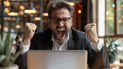 Excited businessman with glasses celebrating success at laptop in modern office setting, fists raised in victorious gesture.