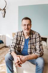 Fototapeta premium Casual mature man wearing glasses and plaid shirt, sitting indoors with a relaxed expression. Comfortable and confident ambiance.