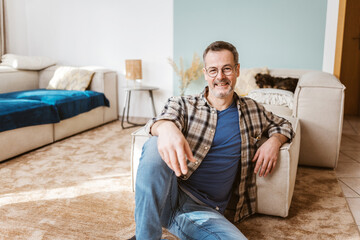 Fototapeta premium A middle-aged man with glasses relaxes on the carpet in his cozy living room, smiling at the camera.