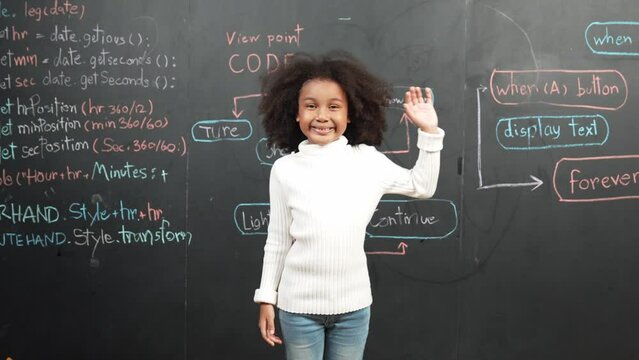 African girl smiling and waving at camera while standing at board with engineering prompt or coding, programing system written in STEM classroom. Child looking at camera while greeting. Erudition.