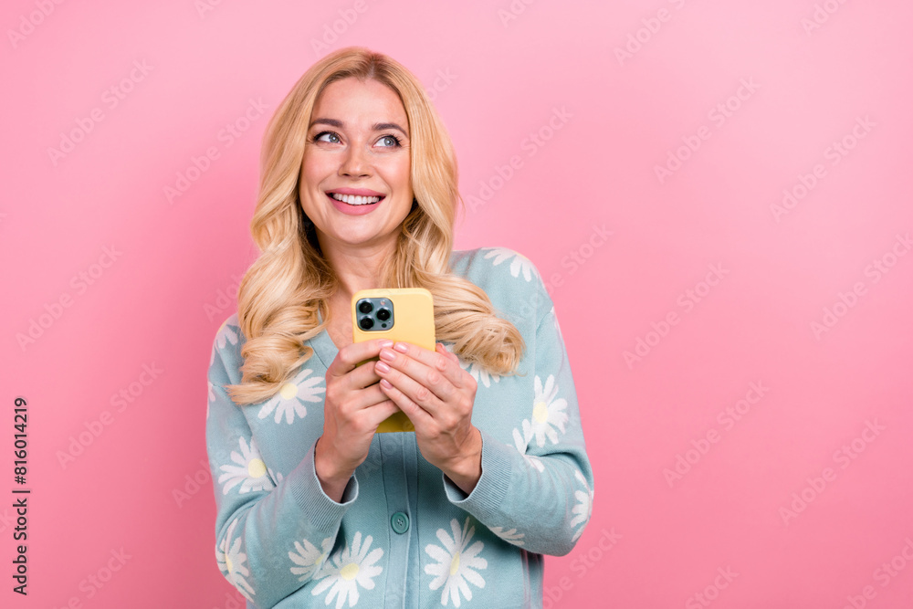 Wall mural Photo of dreamy woman with wavy hairdo dressed print cardigan hold smartphone look at sale empty space isolated on pink color background