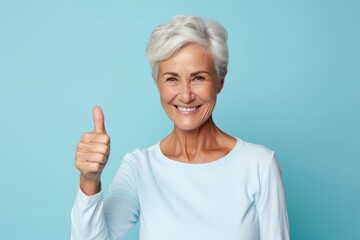 Portrait of a merry woman in her 50s showing a thumb up in soft blue background