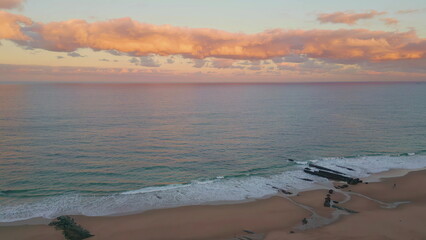 Aerial view dusk waves caressing sandy beach under sunset sky. Pink clouds hover