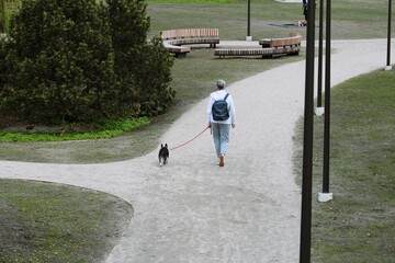 a lady in a white jacket and a black backpack walks through the park with a dog on a leash
