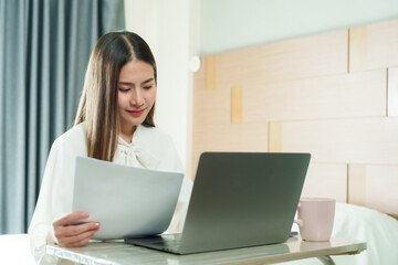 Fototapeta premium Young Asian woman reviewing papers while using laptop in bedroom. Wearing white blouse, long hair. Focused expression, working from home. Modern, comfortable bedroom setting with natural light.