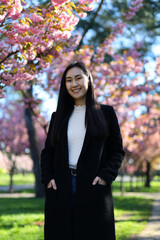 A smiling Korean woman under blooming sakura, exuding happiness in a park portrait. She's attractive.