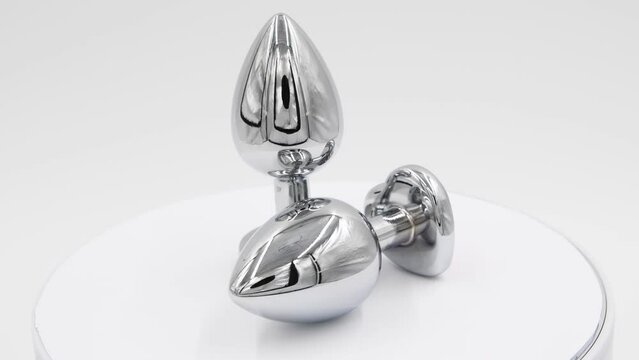 Close-up side view of two metallic butt plugs with pink heart shaped crystals spinning on white background. Soft focus. Real time video. Adult sex toys theme.