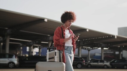 Smiling woman with suitcase standing outdoors, holding phone.