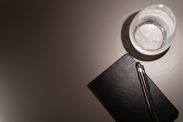 Close up seen from above of small hotel table with black leather diary, pen and glass of water creating contrast of lights and shadows