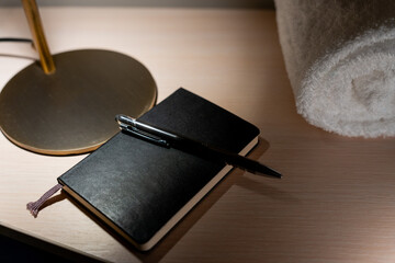 Closeup of hotel room table with lamp illuminating a diary with pen and rolled towel