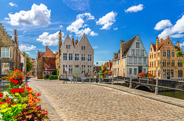 Obraz premium Carmersbrug Bridge stone paved road across water canal, flowers on fence, medieval houses buildings on embankment in Brugge old town, Bruges city historical center, West Flanders province, Belgium