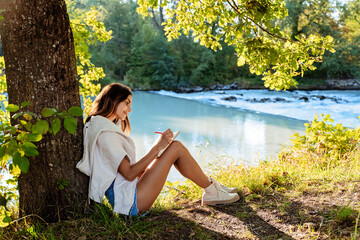 Young woman writing and drawing in notebook sitting under the tree near river on nature in...