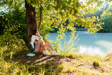 Young happy woman reading a book sitting under the tree near river on nature in sunlight enjoying...