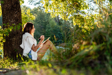 Young attractive girl writing and drawing in notebook sitting under the tree on nature in sunlight....