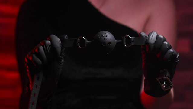 Close-up of female hands in black latex gloves and black dress holding a black BDSM gag on a red background. BDSM - outfit for sex games for adults
