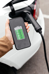 Businesswoman holding smartphone display battery status interface by smart EV mobile application while eco-friendly EV car recharging electricity from charging station