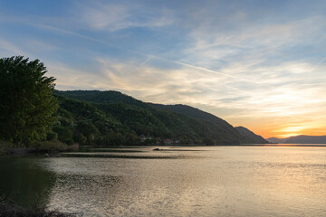 A tranquil lake scene at sunset with calm water, distant mountains, and a silhouette of a village. The sky is painted with orange hues and jet trails, while birds rest on the water's edge