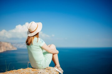 A woman wearing a straw hat is sitting on a rock overlooking the ocean