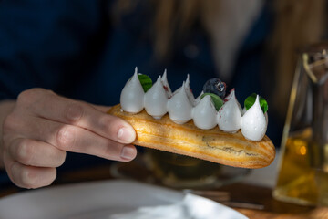 A hand holds a delectable pastry with meringue and cream, garnished with a red dot, on a white plate, with a glass cup and an orange plate with green decorations in the background, under a soft light
