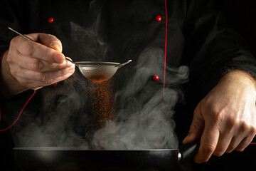 A professional chef adds paprika to a hot frying pan. Concept on a black background of preparing a...