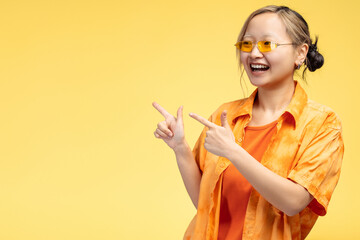 Happy Asian girl in modern glasses pointing to copy space by fingers, isolated on yellow