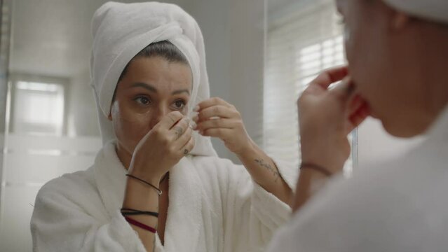 Over the shoulder of Caucasian woman applying under eye patches while taking care of her skin un front of bathroom mirror