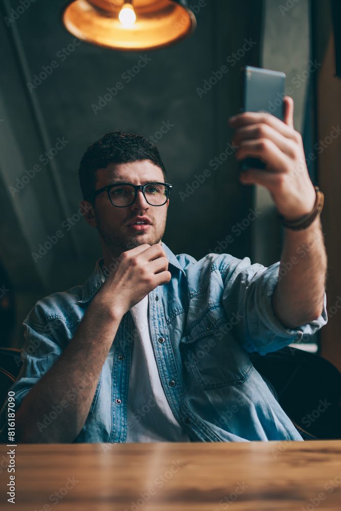 Poster handsome hipster guy in eyeglasses making video conversation with colleague calling by application o
